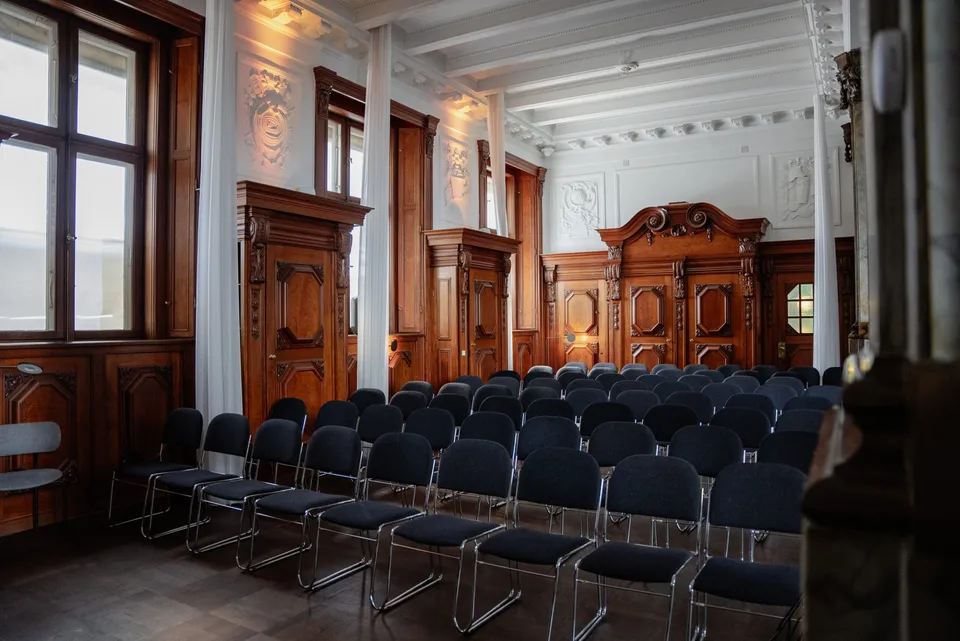 Private Membership Club v. Matrikel1: Klubrummet med stolerækker, høje vinduer og træbeklædte døre.