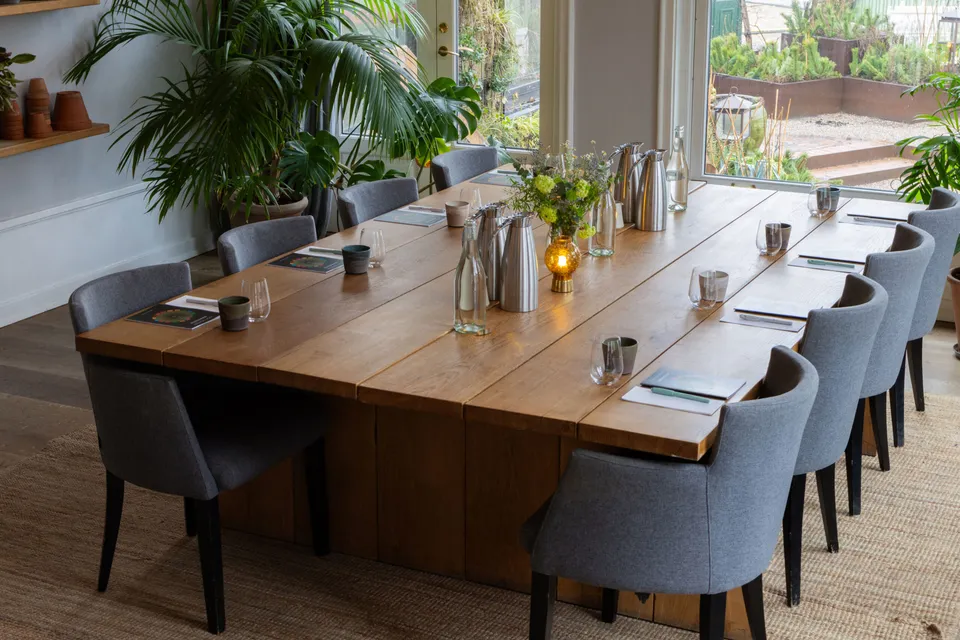 Kabinettet v. Restaurant Kilden: dækket træbord med grå stole og store planter foran et vindue.