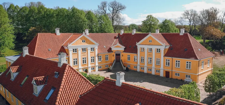 Broløkke Herregård: Herregårdens gule facader og røde tage omkring gårdrummet set fra luften.