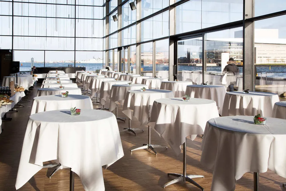 Skuespilhuset v. Det Kongelige Teater: Høje runde borde med hvide duge langs en stor glasfacade mod havnen.