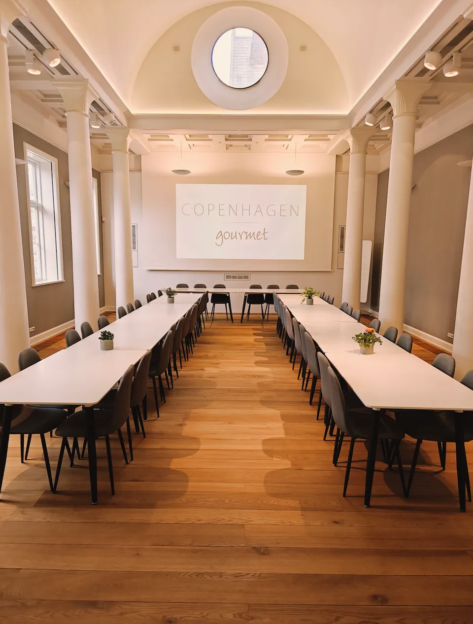 Søjlen v. Copenhagen Gourmet: Det lange lyse rum med bordopstilling, højt loft og skærm.
