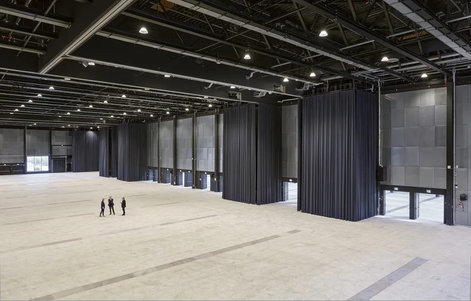 Hal D v. Bella Center Copenhagen: Stor åben hal med højt bjælkeloft, mørke gardiner og glaspartier langs siden.