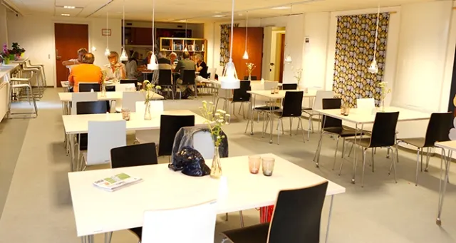 Caféen v. Hyltebjerggård: Lyse caféborde med sorte stole foran en skranke og et åbent køkkenområde.