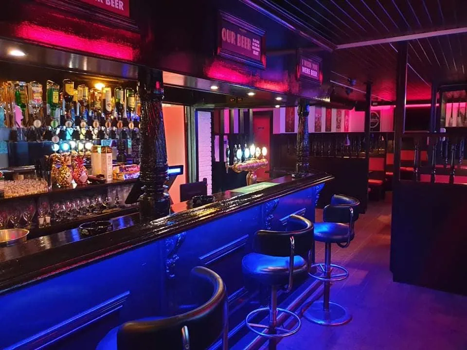 Johnny's Bodega: Bar med høje stole, mange flaskehylder og farvet belysning.