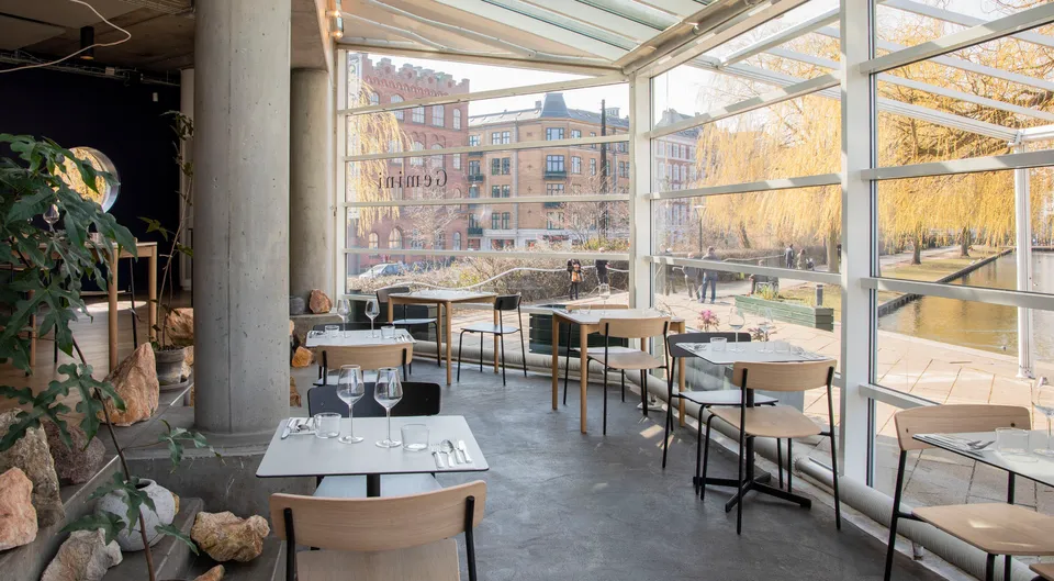Restaurant Gemini: Restaurantborde og træstole i et lyst glasrum ved vandet.