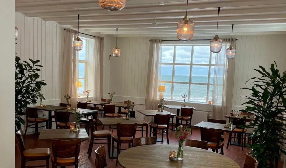 Restauranten v. Gilbjerg Strandhotel: restaurantborde og træstole under synlige bjælker med store vinduer mod vandet.