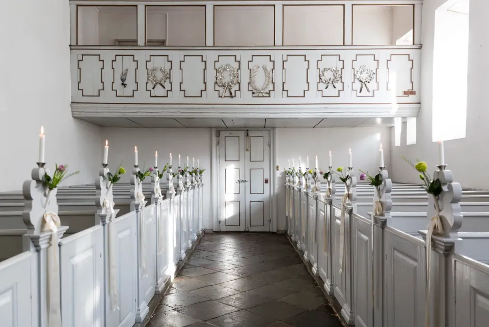 Slotskirken v. Dragsholm Slot: kirkegang mellem hvide bænkerækker med blomster og levende lys.