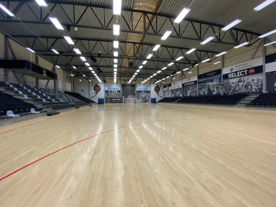 Hal 4 v. Frederiksberghallerne: Sportshal med stort trægulv, mørke tribuner og synlige loftsbjælker.