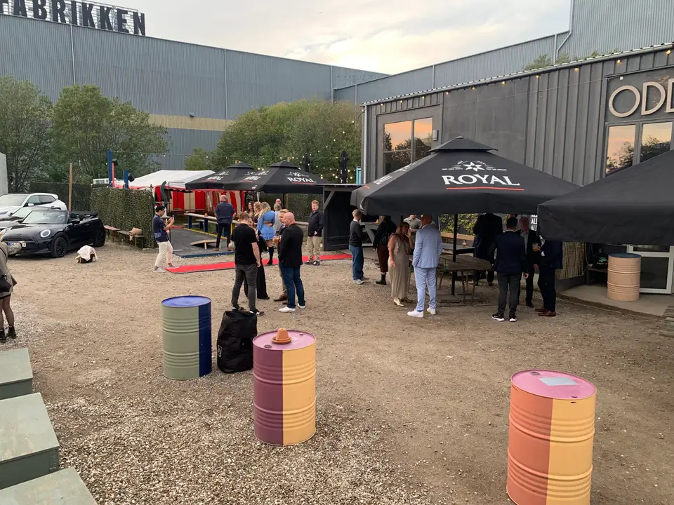 Udeareal og festivalsplads / Odds and Ends: Gæster i en industrigård med parasoller, borde og farvede tønder.
