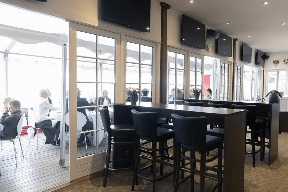 Owners & Trainers Lounge v. Klampenborg Galopbane: Lounge med høje sorte stole og borde langs glasdøre mod en terrasse.