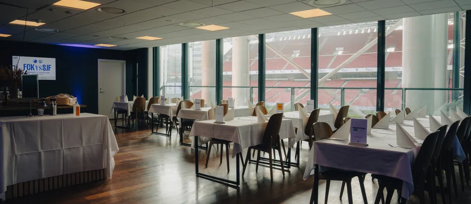 The Balcony v. Parken: dækkede borde i et loungeområde med store vinduer mod stadiontribunen.