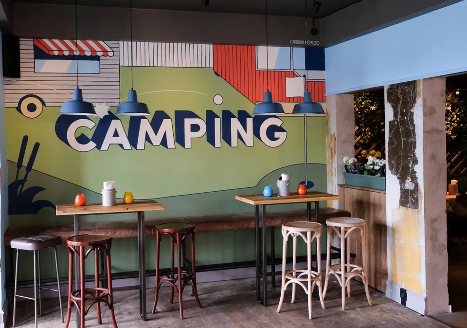 Camping Bar Aaen Aarhus: Høje træborde og barstole ved et farverigt Camping-vægmaleri.