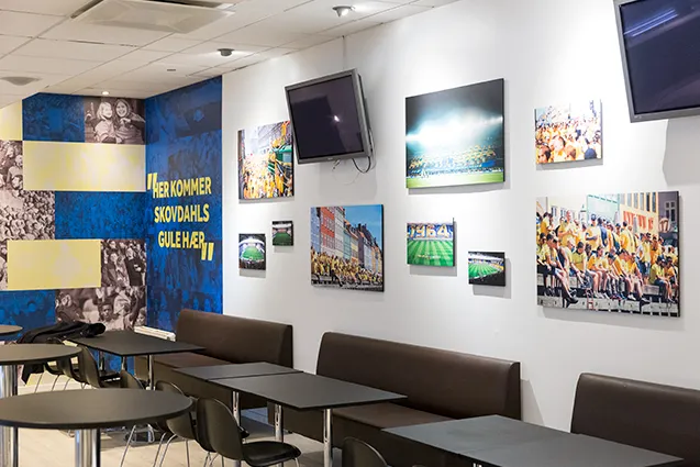 Ebbe Skovdahl Fan Lounge v. Brøndby Stadion: Siddeområde med vægbænke, små borde, skærm og indrammede billeder.