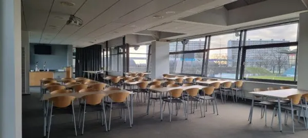 Knud Lundberg Lounge v. Gladsaxe Stadion: borde og træstole i en lounge med store vinduer mod stadiontribunen.