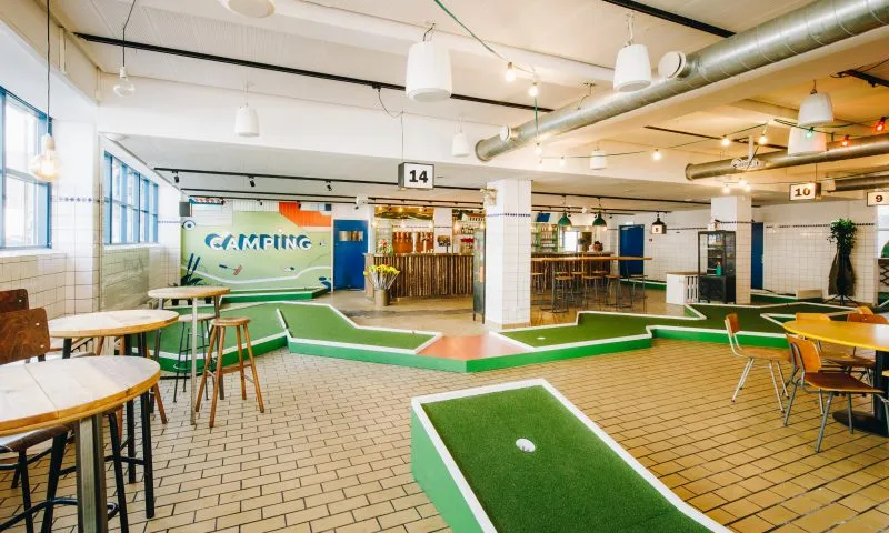 Camping Bar v. Kødbyen: Minigolfbaner og caféborde i et lyst lokale med høje vinduer og en bar.