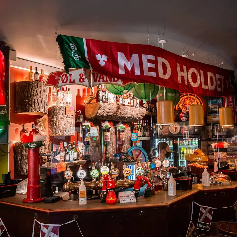 Rævens Bar: Bar med ølhaner, flasker, køleskab og flag over disken.
