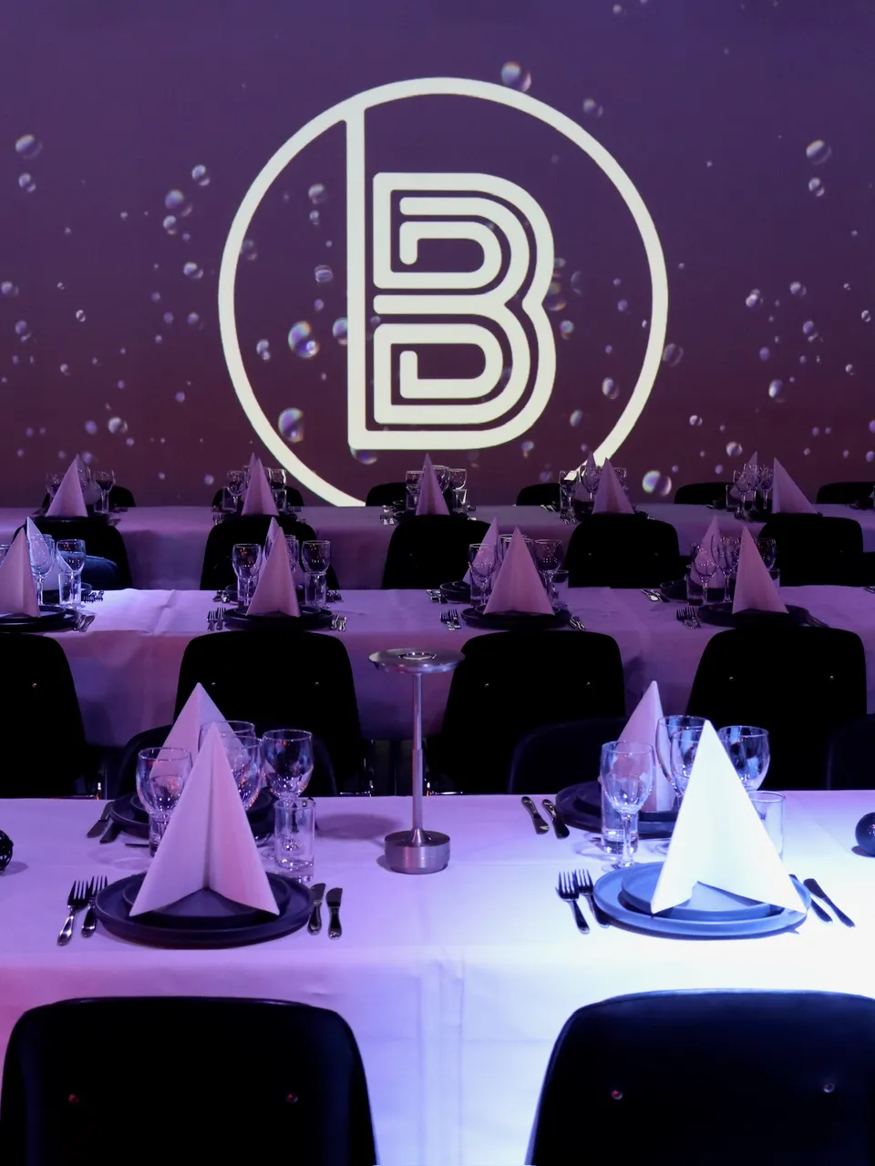 The Ballroom Aarhus: dækkede bordrækker med hvide duge og lilla lys foran en vægprojektion.