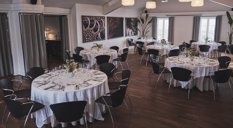 Førstesalen v. Restaurant Sletten: runde dækkede borde i et lyst lokale med mørkt trægulv og kunst på væggene.