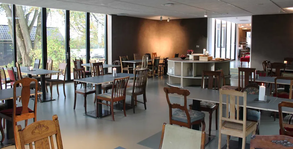 Cafeen v. Kirstinelund: Lyst caféområde med træstole, borde og store vinduer.