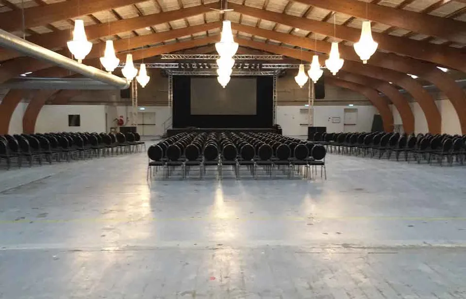 Hallen v. Savværket: Stor hal med buede træbjælker, stoleopstilling og scene.